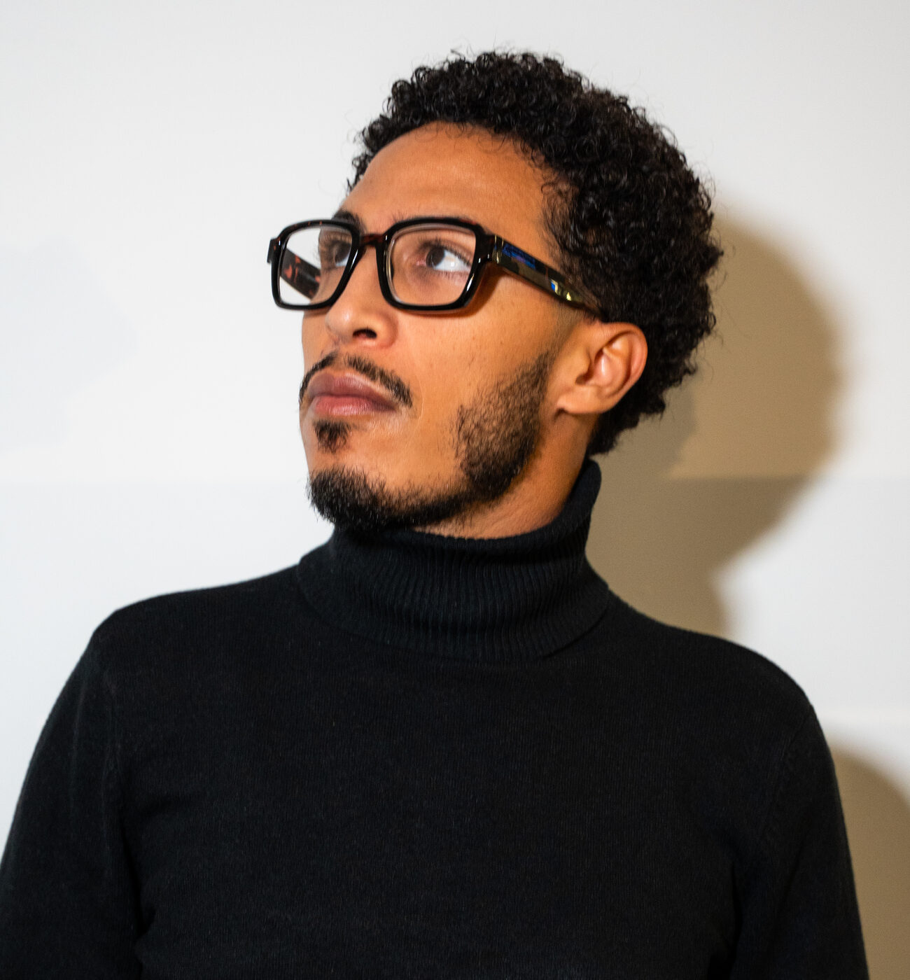 Hamza Bouzhar in glasses and a black turtleneck, looking up and to his right against a white wall.